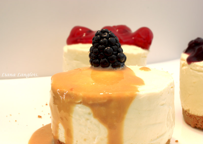 Cheesecake A close up photo of two individual cheesecake desserts, one with dulce de leche sauce and a blackberry, and the other with cherry topping.
