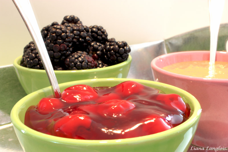 Cheesecake A close up photo of 2 sauces (cherry topping and dulce de leche) in one green bowl, and one pink, as well as a green bowl of blackberries.