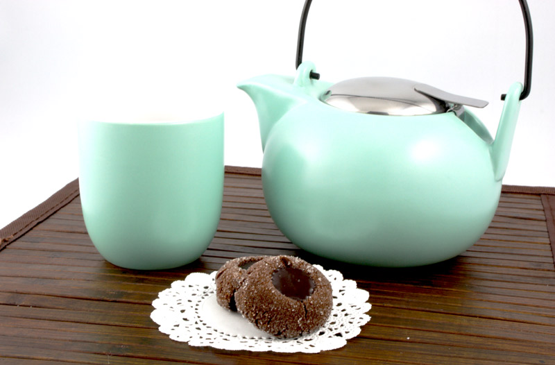 A photo of 2 chocolate thumbprints on a white doilie with a turquoise chinese tea pot and teacup on a brown wooden placemat.
