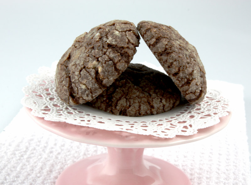 A close up photo of 3 dark chocolate-almond crackles on a mini pink cake stand on a white tea towel.