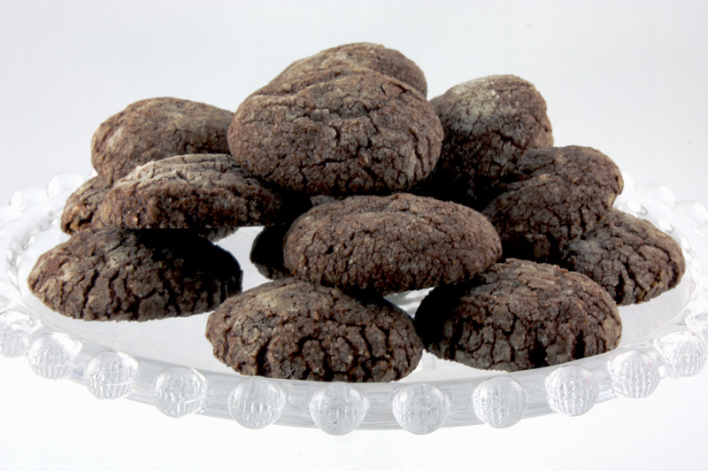 A close up photo of dark chocolate-almond crackles on a glass cake stand.
