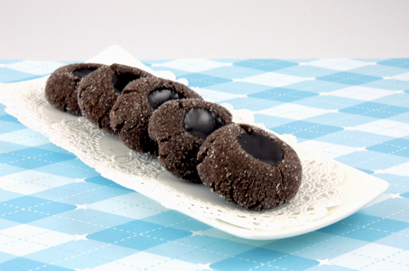 A close up photo of chocolate thumbprints on a white dish with white doilie on a blue patterned sheet of paper.