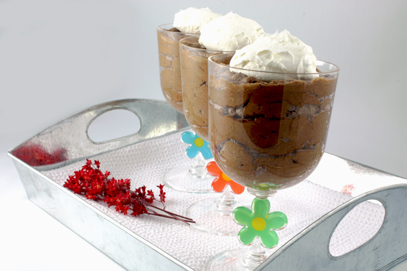 Gordon Ramsay Four-Minute Chocolate Mousse A close up photo of three chocolate mousse desserts in clear wine tumblers on a tin tray lined with a white waffle pattern tea towel, with a sprig of fresh red flowers.