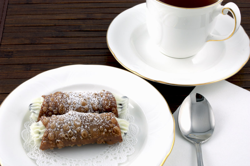 A close up photo of a teacup and saucer, a silver spoon on a white napkin and cannoli siciliani served on a white plate lined with a white doilie all on a dark wooden placemat.