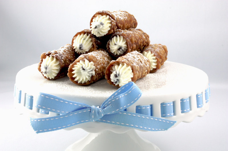 A photo of Cannoli Siciliani (Italian pastries) on a white cake plate with a blue and white ribbon.