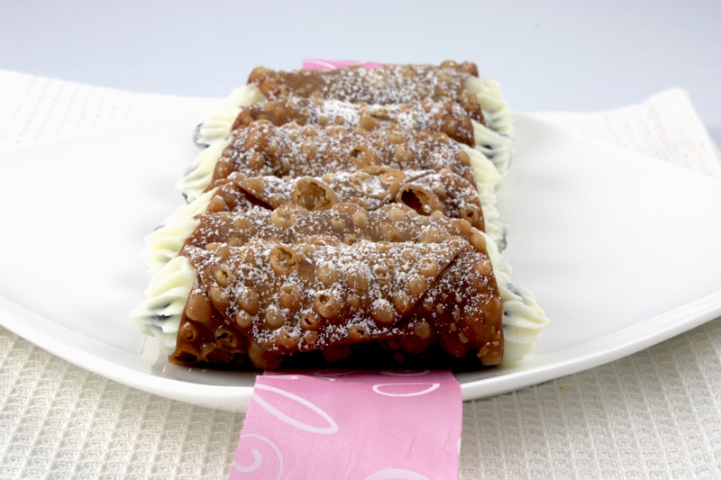 A photo of Italian, Cannoli Siciliani pastries plated on a white plate with a pink and white ribbon placed on top of a white, waffle patterned tea towel.