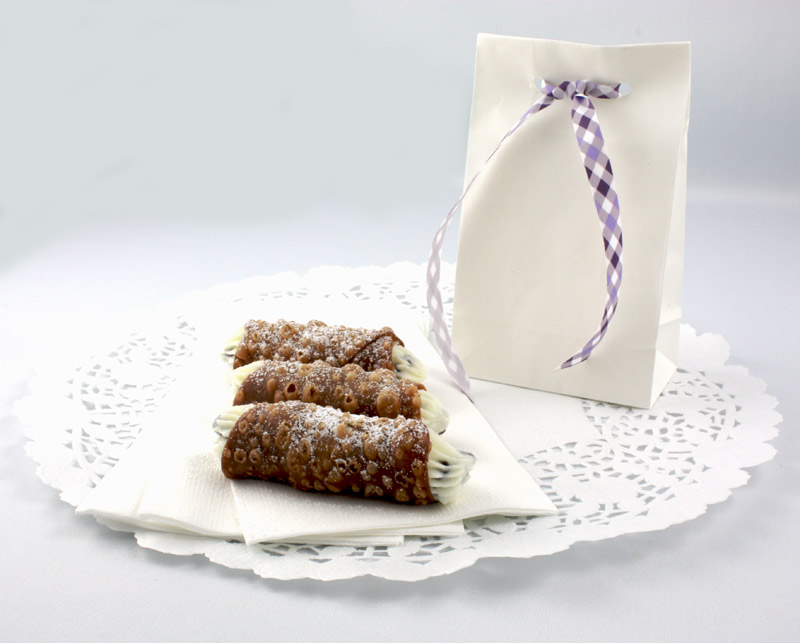 A photo of Cannoli Siciliani (Italian pastries) sitting on three white napkins on a large white doilie along side a white bag with a purple tied ribbon.