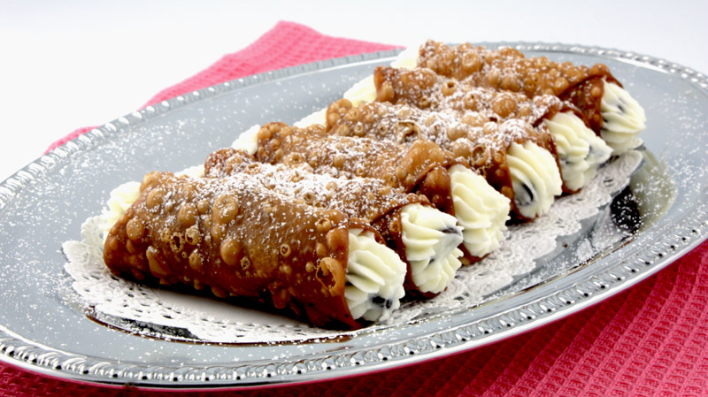 A close up photo of Cannoli Siciliani (Italian Pastry) on a silver tray lined with white doilies on a pink placemat.