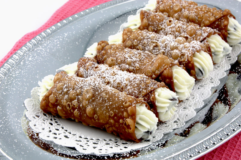 A close up photo of Cannoli Siciliani (Italian Pastries) on a silver serving tray lined with small white doilies sitting on top of a pink, waffle patterned, tea towel.