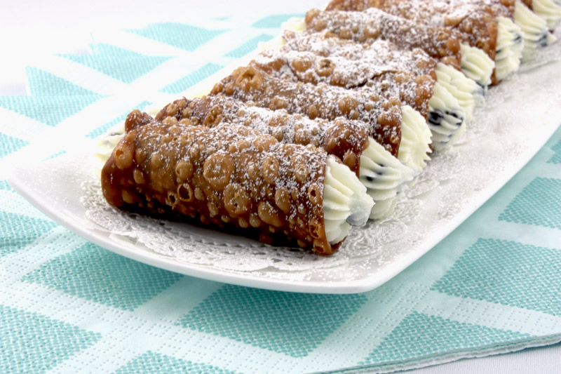 A close up photo of Cannoli Siciliani (Italian pastries) on a white plate lined with white doilies placed on a bed of blue and white napkins.