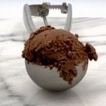 A photo of a scoop of chocolate ice cream in a silver ice cream scoop dispenser on a marble slab.