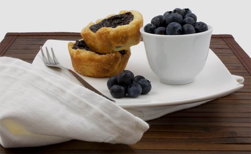 Chocolate Custard Tartlettes A photo of 2 chocolate custard tartlettes served with a bowl of blueberries on a white plate with a silver fork and a white napkin placed on a wooden placemat.