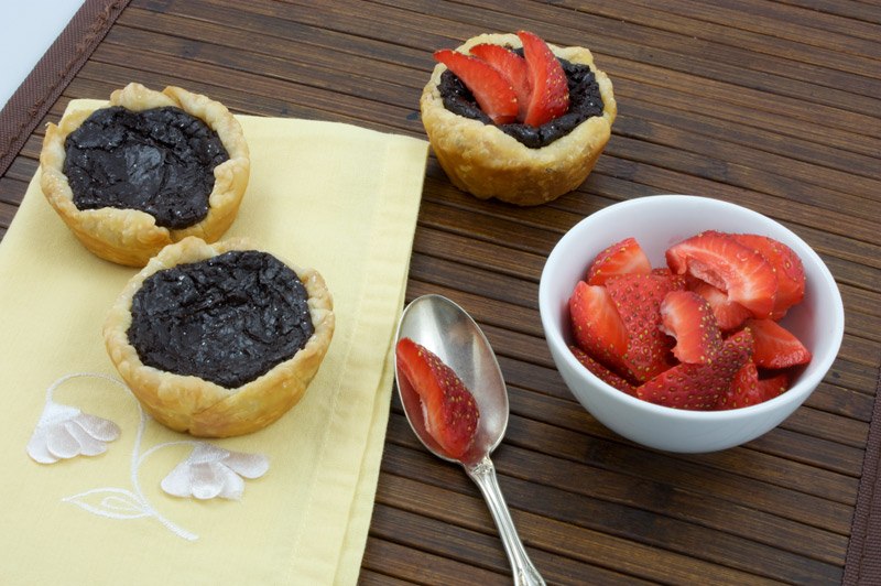 Chocolate Custard Tartlettes A photo of 2 chocolate custard tartlettes on a yellow napkin along side a silver spoon, a bowl of strawberries and a chocolate custard tartlette with strawberries on top placed on a wooden placemat.