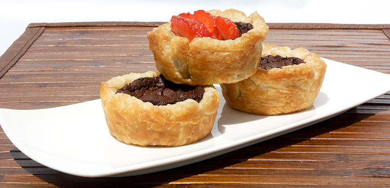 Chocolate Custard Tartlettes A photo of 3 chocolate custard tartlettes, one with strawberries, placed on a white dish on a wooden placemat.