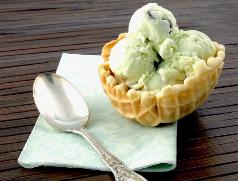 Mint Chocolate Chip Ice Cream A photo of mint chocolate chip ice cream in a waffle bowl on a green napkin with a silver spoon on a wooden placemat.
