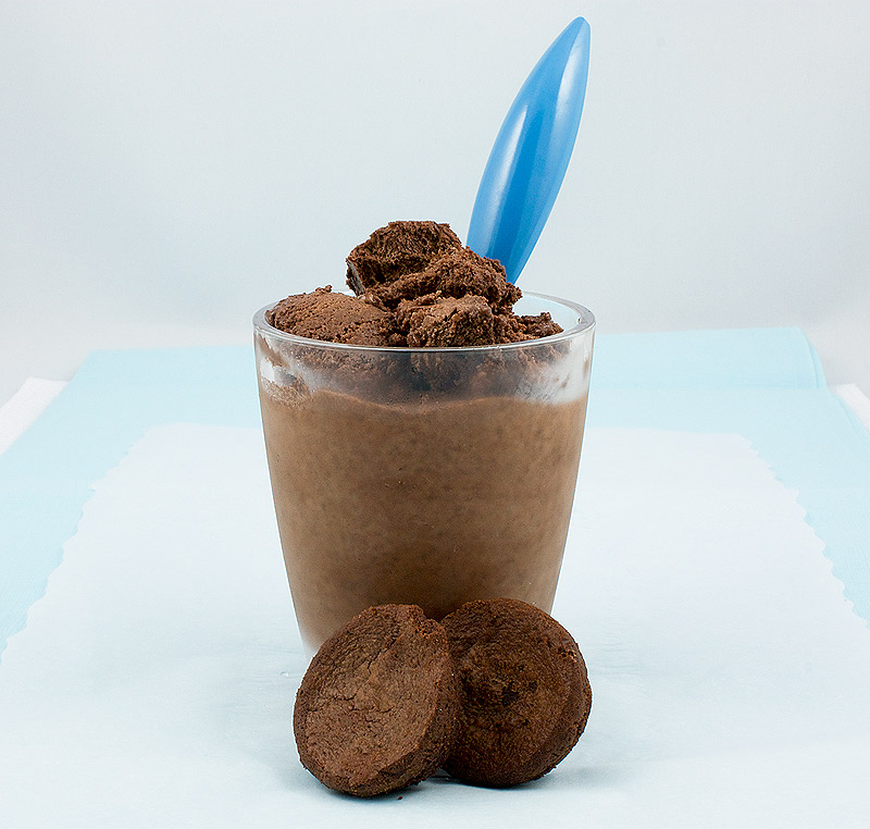 A photo of a cup of double chocolate brownie semifreddo garnished with chopped brownies, with a blue spoon inserted in cup and 2 small brownie bites displayed in front of glass on a piece of white parchment that is sitting atop a blue napkin.