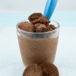 A close up photo of a double chocolate brownie semifreddo dessert served in a glass and garnished with brownie bits, a blue spoon is inserted in glass and 2 brownie bites are placed in front of glass.