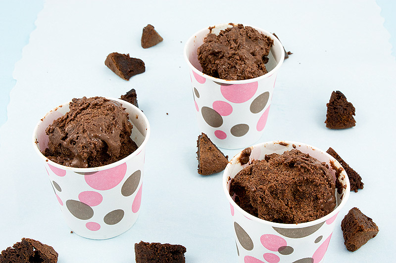 A close up photo of 3 double chocolate brownie semifreddo desserts in pink and brown polka dotted paper cups, sitting on a piece of parchment with small brownies bites scattered around the cups.