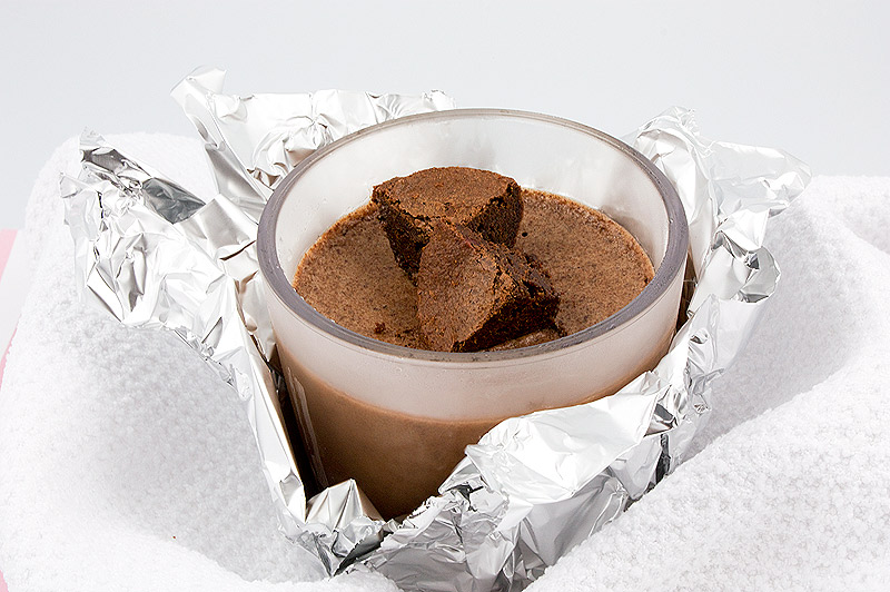 A close up photo of a double chocolate brownie semifreddo dessert served in a clear tumbler garnished with brownie bites, tumbler is wrapped in tin foil, nestled in a white tea towel.
