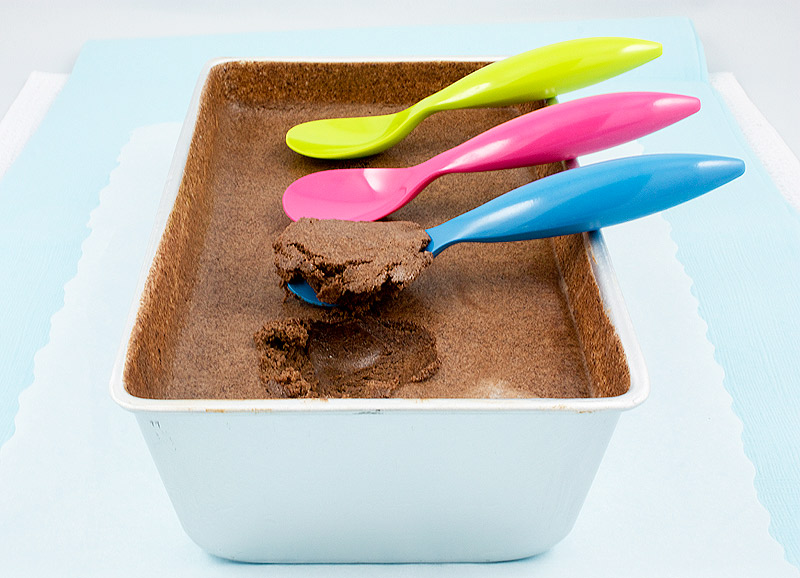 A photo of Donna Hay's double chocolate semifreddo in a tin baking dish with three plastic spoons, one blue, one pink and one yellow, arranged in a row, one with a scoop of semifreddo.