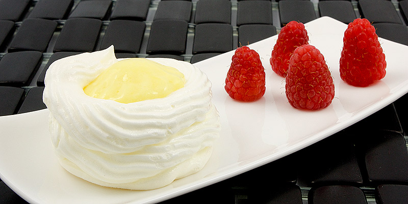 lemon curd meringue nests, meringue nests, meringue cups, easy lemon curd A photo of a lemon curd meringue nest on a white plate with raspberries on the side, placed on a black placemat.