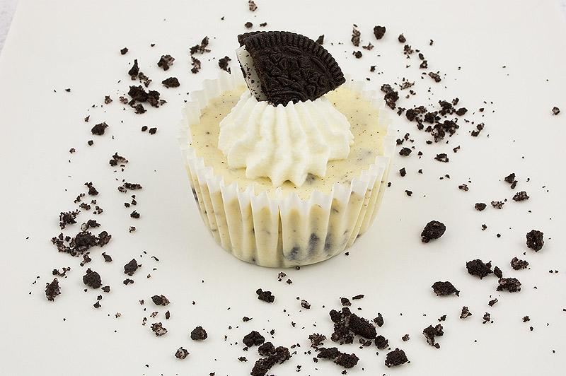 Oreo cookies and cream cheesecakes, Martha Stewart's cookies and cream cheesecakes recipe, easy recipe for Oreo cookies and cream cheesecake cupcakes A photo of an Oreo cookies and cream cheesecake with whipped cream and an Oreo cookie on top, placed on a white plate with Oreo cookie crumbs scattered on the dish.
