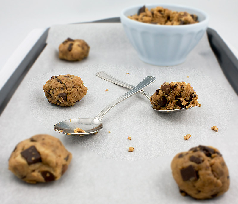 how to make the best chocolate chip cookies, secret to best chocolate chip cookies, secret to the best chocolate chip cookies A photo of a bowl of Jacques Torres's Secret Chocolate Chip Cookie dough in a pale blue bowl with 2 silver spoons, one containing a small portion of cookie dough and a few scoops of cookie dough proportioned and prepared to bake placed on a cookie sheet lined with parchment paper.