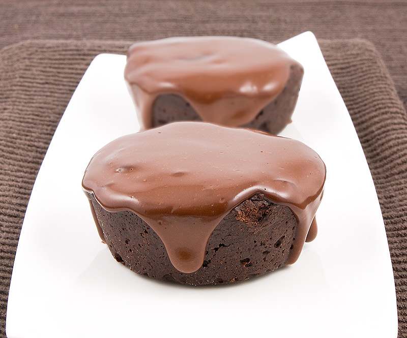 A close up photo of 2 flourless chocolate cakes with chocolate glaze on a white, rectangular plate resting on a chocolate brown tea towel.