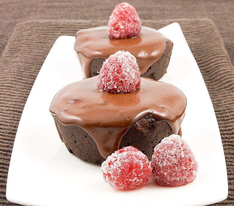A photo of 2 flourless chocolate cakes with chocolate glaze, garnished with sugared raspberries on a white rectangular plate placed on a chocolate brown tea towel.