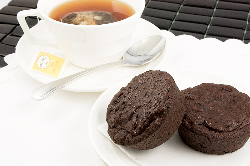 A close up photo of 2 flourless chocolate cakes on a white dish served with a cup of tea in a white teacup with saucer and silver spoon placed on a white napkin resting on a black placemat.