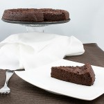 David Lebovitz desserts, french dessert recipes, flourless chocolate torte A photo of a chocolate flourless cake on a cake stand with a slice of flourless chocolate cake placed in front on a white, square plate with a fork.