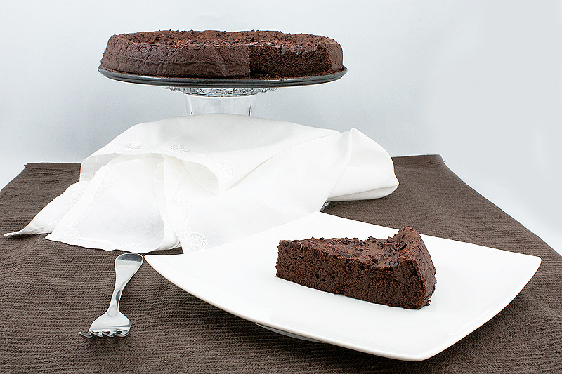 simple dessert recipes, flourless cake recipe, easy chocolate cake recipes A photo of David Lebovitz's Racines Cake on a glass cake stand placed on a brown tea towel and also a slice of the dessert placed on a square, white plate with a dessert fork and white, cloth napkin.