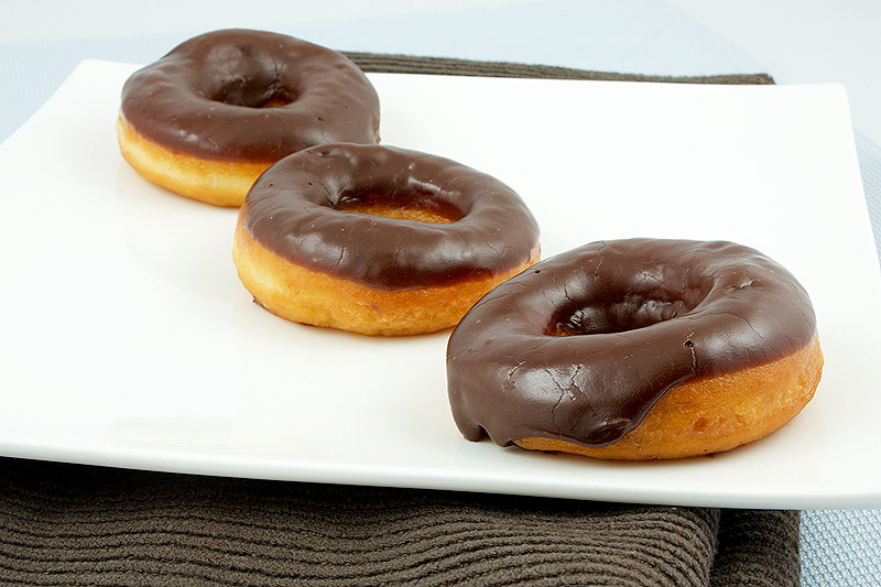 how to make doughnuts at home, doughnut recipe, chocolate doughnut glaze A photo of 3 chocolate glazed doughnuts on a white plate placed on a brown tea towel.