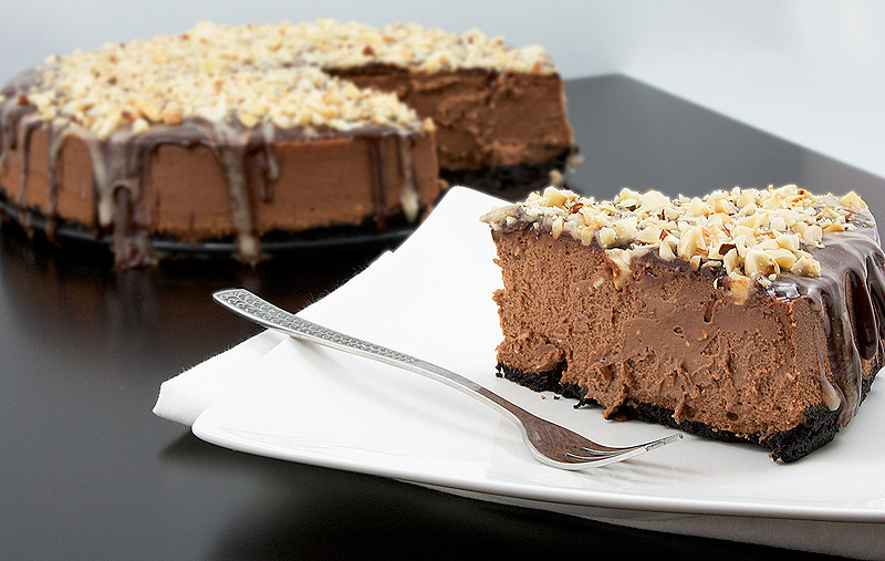 chocolate cheesecake recipe, simple chocolate dessert, easy dessert recipes A photo of a slice of chocolate cheesecake with Oreo cookie crust topped with chocolate sauce, butter caramel sauce and toasted hazelnuts on a white dish with a dessert fork and full cheesecake placed in behind.