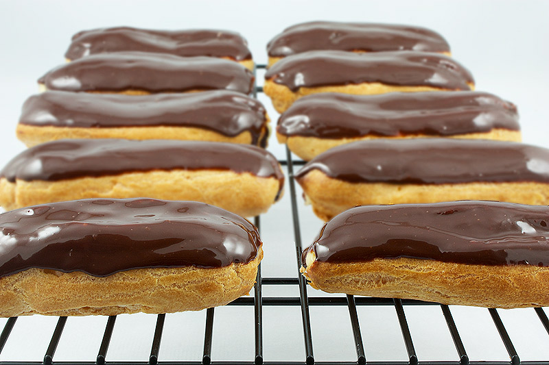 Chocolate Éclairs with Vanilla Bean Crème Pâtissière and Chocolate Ganache Glaze A close up photo of Chocolate Éclairs with Vanilla Bean Crème Pâtissière and Chocolate Ganache Glaze french pastries on a cooling rack.