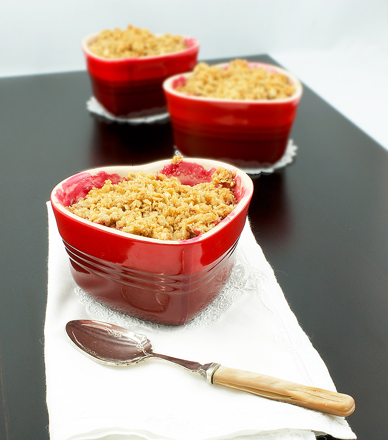 fruit crumble, quick and easy desserts, rhubarb crumble recipe A photo of a Pear, Rhubarb and Raspberry Fruit Crumble dessert in a red, heart-shaped ramekin placed on a white napkin with a dessert spoon and two additional fruit crumbles placed in behind.