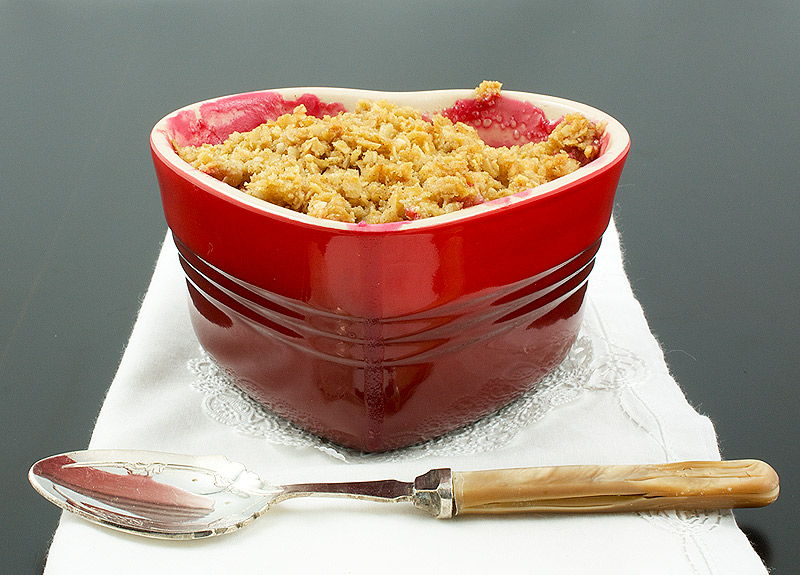 fruit crisp recipe, simple fruit dessert, crumble toppings A photo of a Pear, Rhubarb and Raspberry Fruit Crumble dessert in a red, heart-shaped ramekin placed on a white napkin with a dessert spoon.