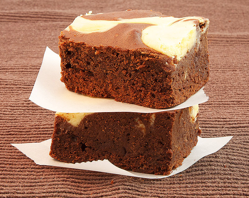 A photo of 2 Baileys® Cheesecake Swirl Chocolate Brownie squares, stacked with pieces of parchment separating the squares.