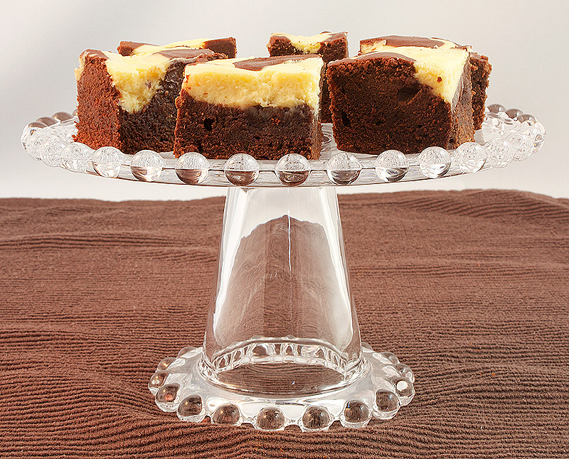 A photo of Baileys® Cheesecake Swirl Chocolate Brownies on a glass cake stand.