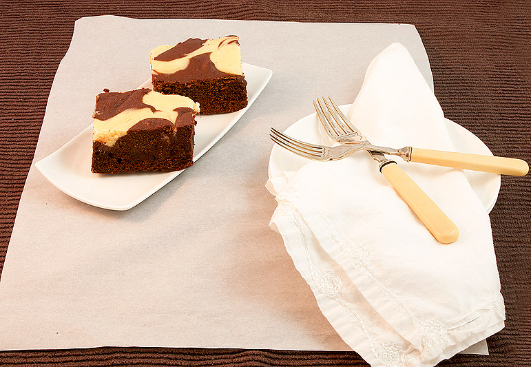 A photo of 2 Baileys® Cheesecake Swirl Chocolate Brownie squares on a small, rectangular white plate along side 2 dessert plates with a cloth napkin and 2 dessert forks.