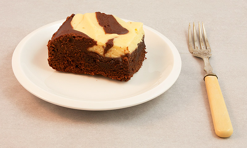A photo of a Baileys® Cheesecake Swirl Chocolate Brownie square on a small white plate with a dessert fork.