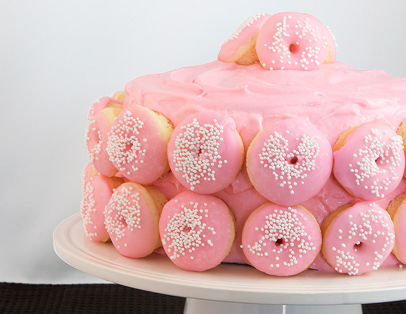 homemade cream cheese frosting, how to make cream cheese frosting, cream cheese frosting recipes A close up photo of Ina Garten's Chocolate Layer Cake with White Chocolate Cream Cheese Frosting and decorated with mini Baked White Chocolate Doughnuts with pink tinted Vanilla Doughnut Glaze and sprinkled with white nonpareils on a white cake stand.