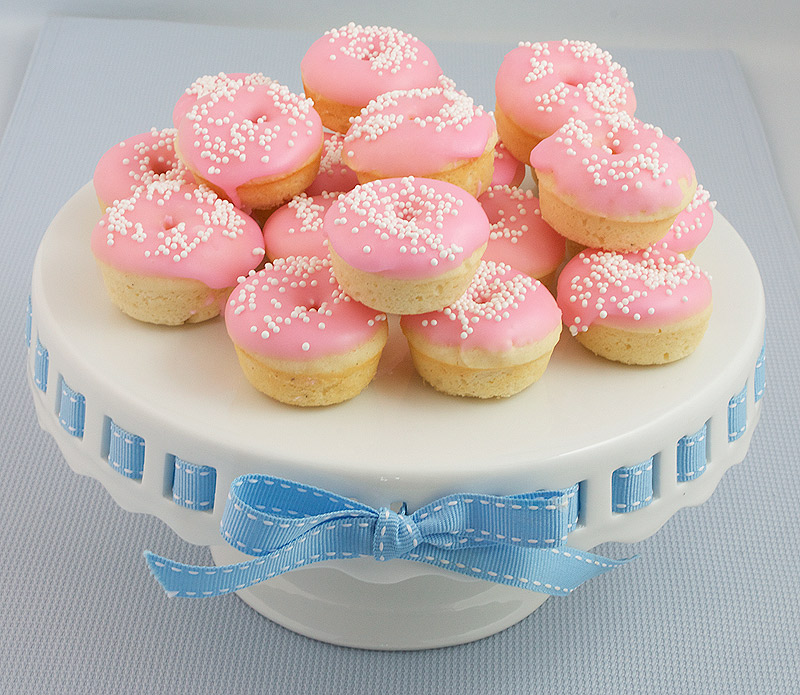 A photo of Baked White Chocolate Cake Doughnuts with pink Vanilla Doughnut Glaze decorated with white nonpareils on a white cake stand.