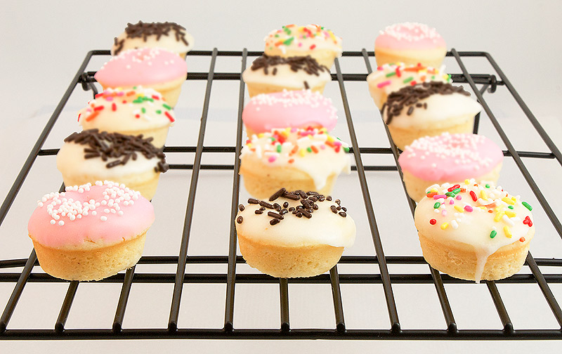 A photo of Baked White Chocolate Cake Doughnuts with Vanilla Glaze and decorated with sprinkles on a wire rack.