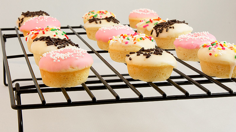 A photo of mini baked cake doughnuts with vanilla doughnut glaze and decorated with assorted sprinkles cooling on a wire rack.