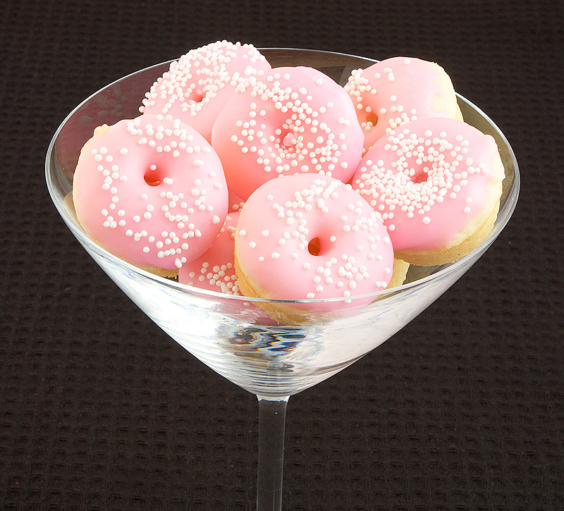 A photo of Baked White Chocolate Cake Doughnuts with pink Vanilla Doughnut Glaze and decorated with white nonpareils in a clear martini glass.