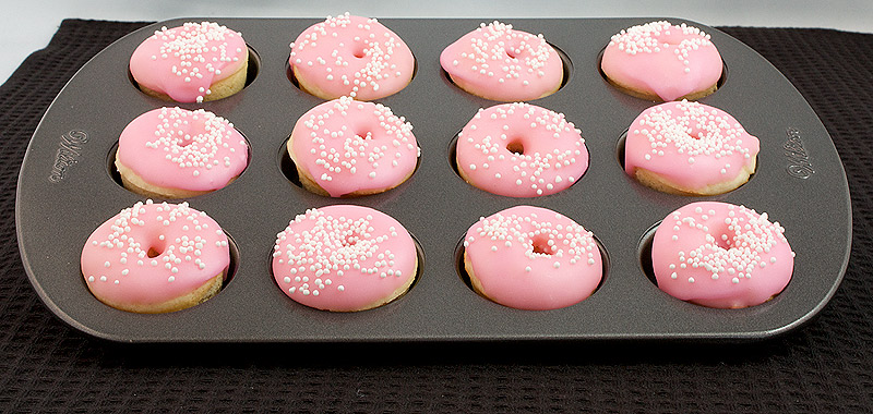 A photo of mini doughnuts with pink doughnut glaze and decorated with white nonpareils in a Wilton mini doughnut pan.