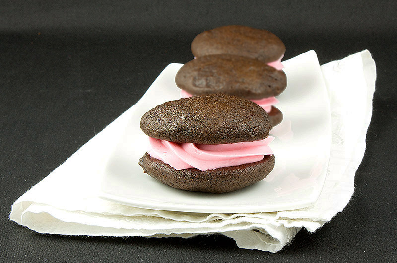 Chocolate Whoopie Pies A photo of 3 Chocolate Whoopie Pies with Pink Vanilla Swiss Meringue Buttercream filling placed in a row on a white rectangular plate.