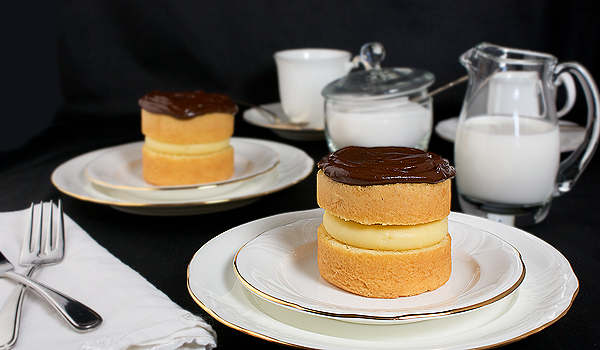 Mini Boston Cream Pies A photo of two Mini Boston Cream Pies desserts shown along side 2 coffee cups with saucers and glass cream filled jug and sugar bowl.