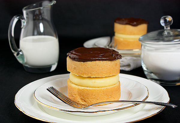 Boston Cream Pies A photo of Boston Cream pies served in mini form on white dessert dishes shown with a glass creamer and glass sugar bowl.